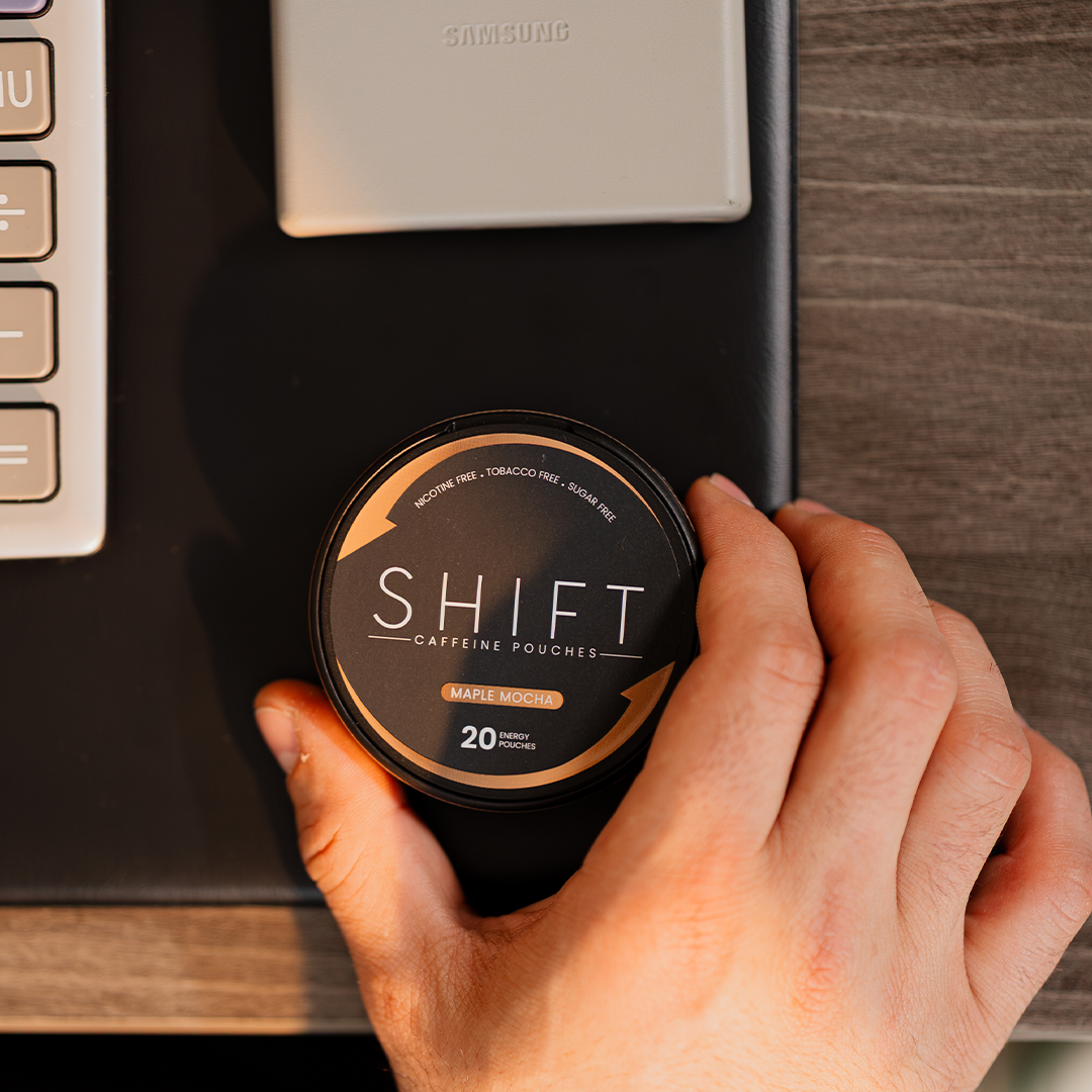 Hand holding a container of 'SHIFT' caffeine pouches on a desk with a keyboard in the background.